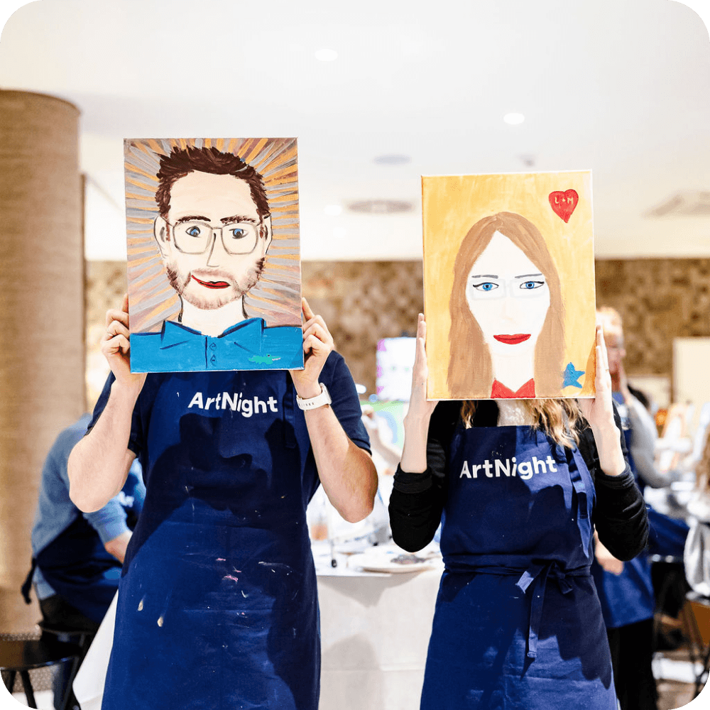ArtNight Paint your Partner Zwei Personen halten bemalte Porträts vor ihre Gesichter. Beide tragen in einem hell erleuchteten Raum violette Schürzen mit der Aufschrift „ArtNight“.
