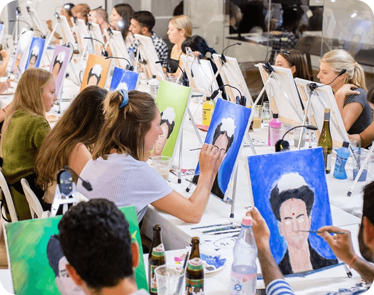 ArtNight Team & Firmenevents Menschen bei einer ArtNight, die Porträts auf Staffeleien malen, mit verschiedenen bunten Leinwänden und Malutensilien auf den Tischen.