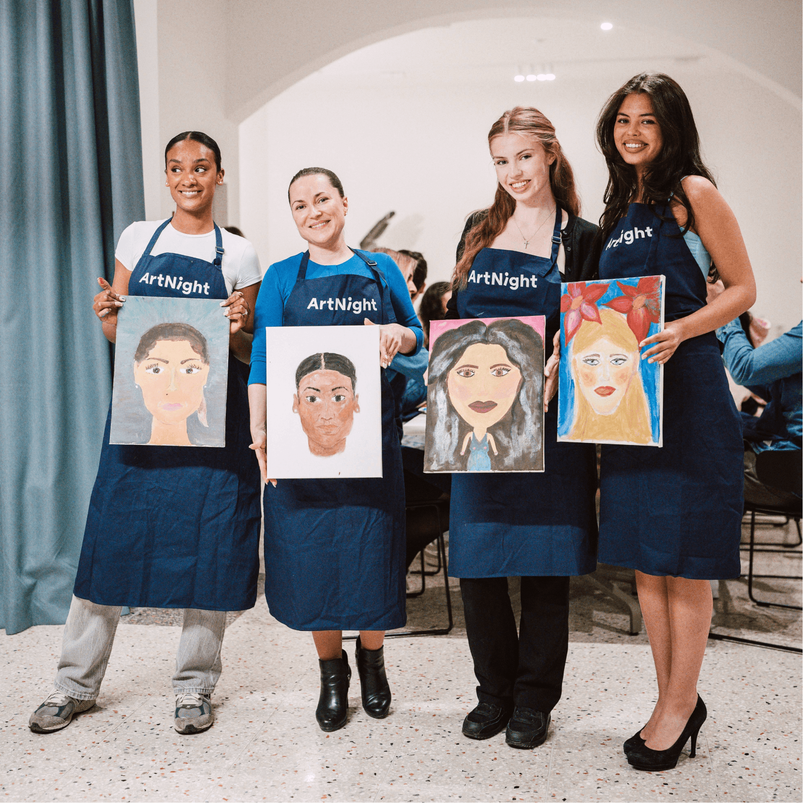 ArtNight Paint your Partner Vier Freunde, die „ArtNight“-Schürzen tragen, präsentieren Porträtgemälde und lächeln in einem Innenraum.