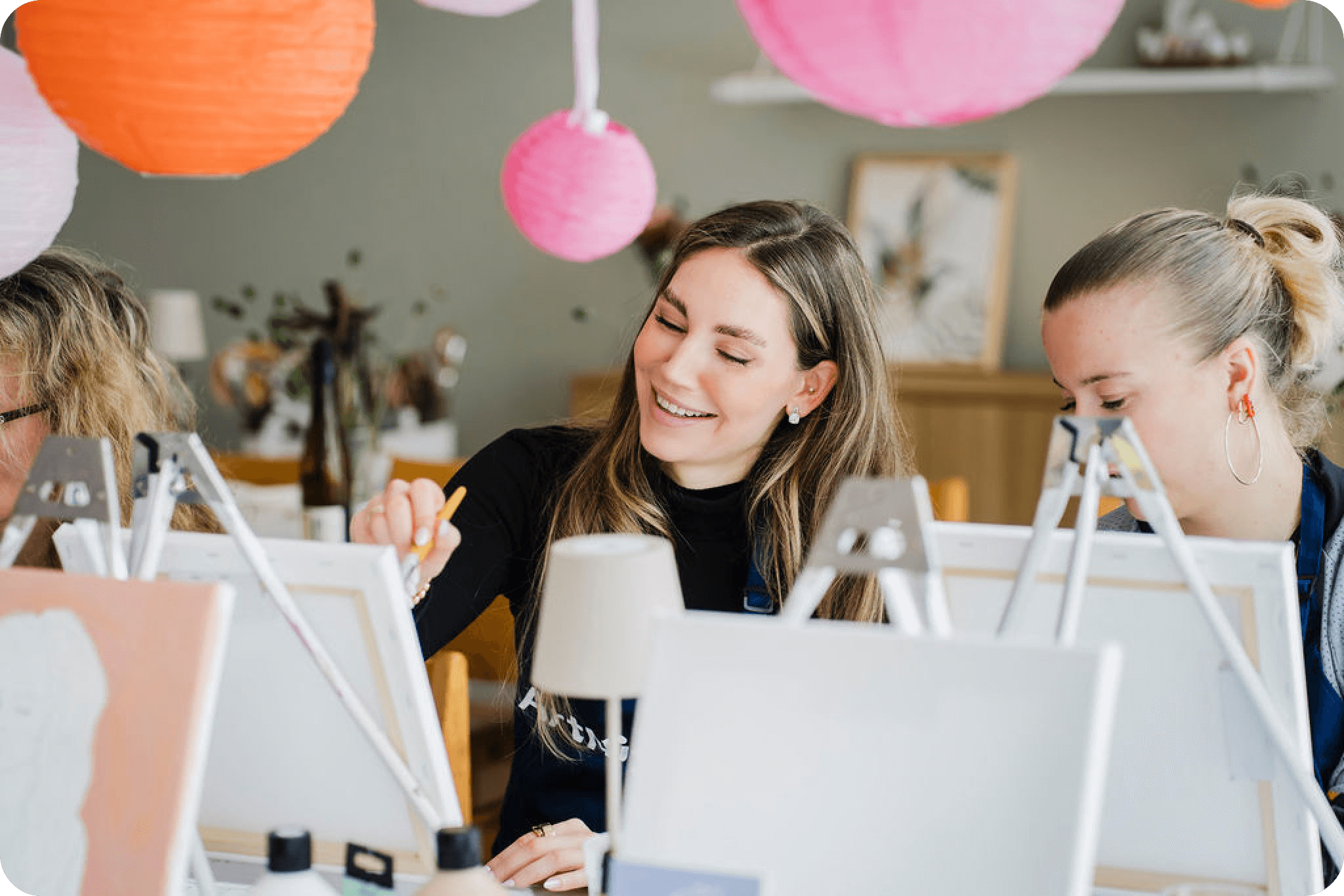 ArtNight Kreativ JGA Drei Frauen malen auf Leinwänden in einem mit bunten Papierlaternen geschmückten Atelier, wobei eine von ihnen lächelt und sich mit ihrem Kunstwerk beschäftigt.