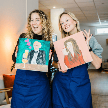 ArtNight Team Events Zwei lächelnde Menschen, die bunte Gemälde in den Händen halten. Sie tragen ArtNight-Schürzen und stehen in einem hellen Raum mit gemütlicher Atmosphäre.