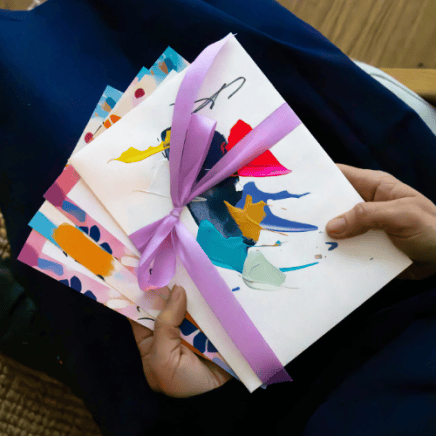 ArtNight malkurs gutschein Hände, die bunte abstrakte Kunstwerke halten, die mit einem violetten Band zusammengebunden sind, vor einem marineblauen Hintergrund.