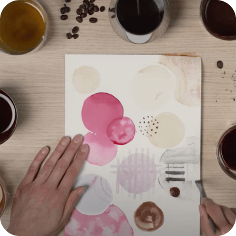 AN Masterclass Hände malen mit Kaffee auf Papier, umgeben von Kaffeetassen in verschiedenen Farbtönen auf einem Holztisch.