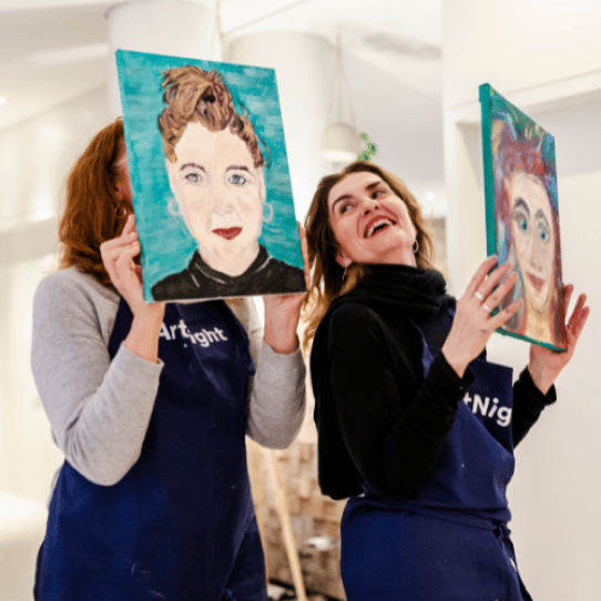 Paint your Partner Zwei Personen in ArtNight-Schürzen halten in einem hell erleuchteten Raum Porträtgemälde hoch, lächeln und präsentieren ihre Kunstwerke.