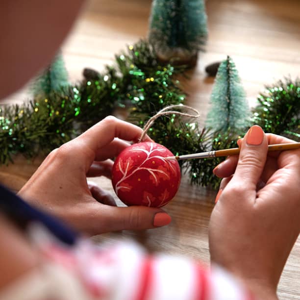 weihnachtskugeln Person, die mit einem Pinsel ein rotes Weihnachtsornament bemalt, umgeben von kleinen Bäumen und grünem Lametta auf einem Holztisch.