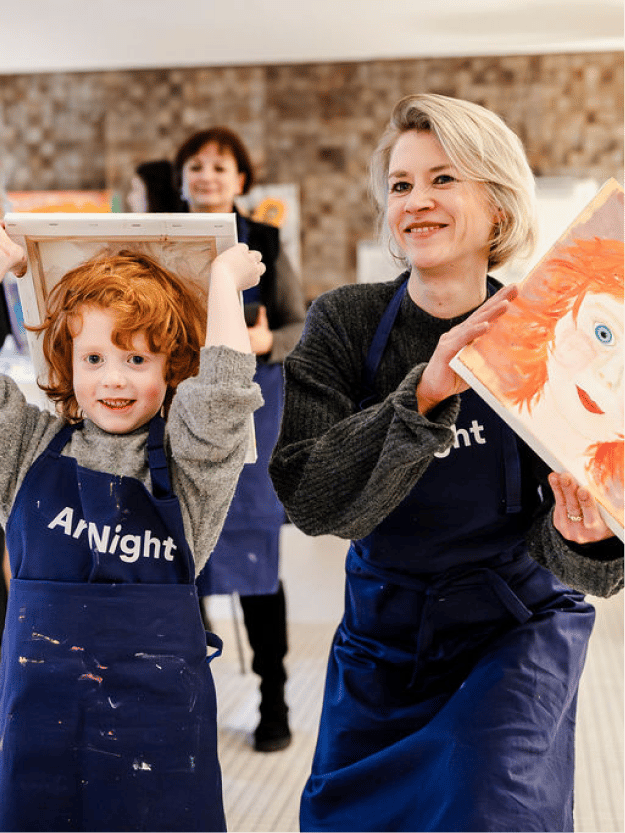 Paint your Family Ein Kind und ein Erwachsener, beide mit ArtNight-Schürzen bekleidet, präsentieren während eines Kunstkurses freudig ihre bemalten Leinwände.