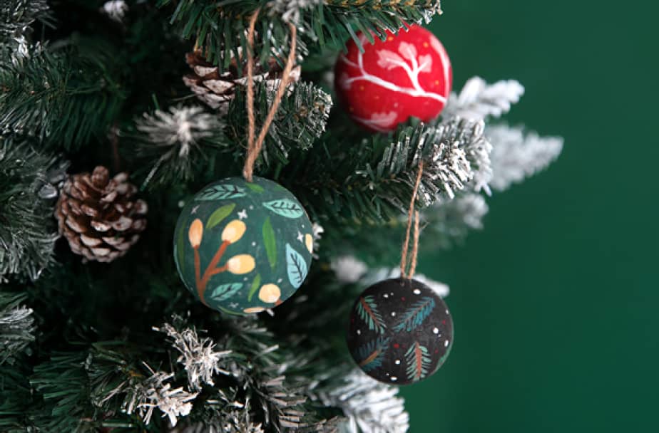 ArtNight Weihnachten Weihnachtsbaum mit schneebedeckten Zweigen, Tannenzapfen und drei handbemalten Ornamenten in Rot, Grün und Schwarz mit festlichen Motiven.