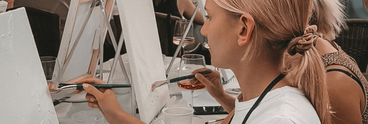 Eine Person mit blondem Haar, die in einer Gruppe auf einer Leinwand malt, hält einen Pinsel und ein Glas.