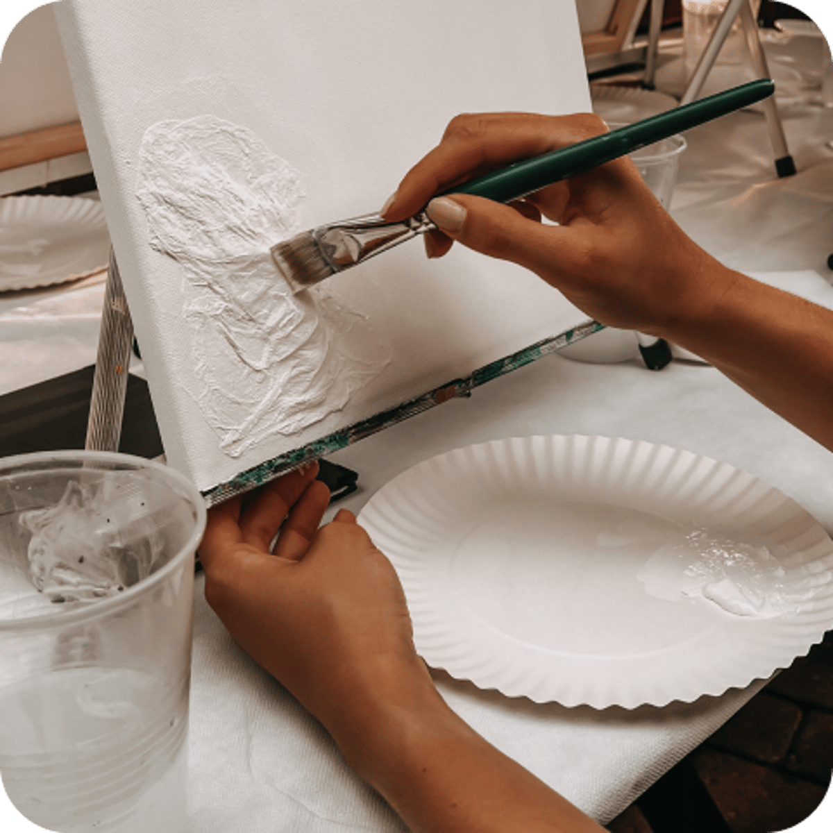 Eine Person malt mit einem Pinsel strukturiertes Weiß auf eine Leinwand, umgeben von Papptellern und einem Plastikbecher auf einem Tisch.