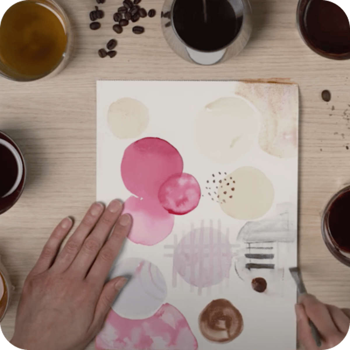 Hände malen mit Kaffee auf Papier, umgeben von Kaffeetassen in verschiedenen Farbtönen auf einem Holztisch.