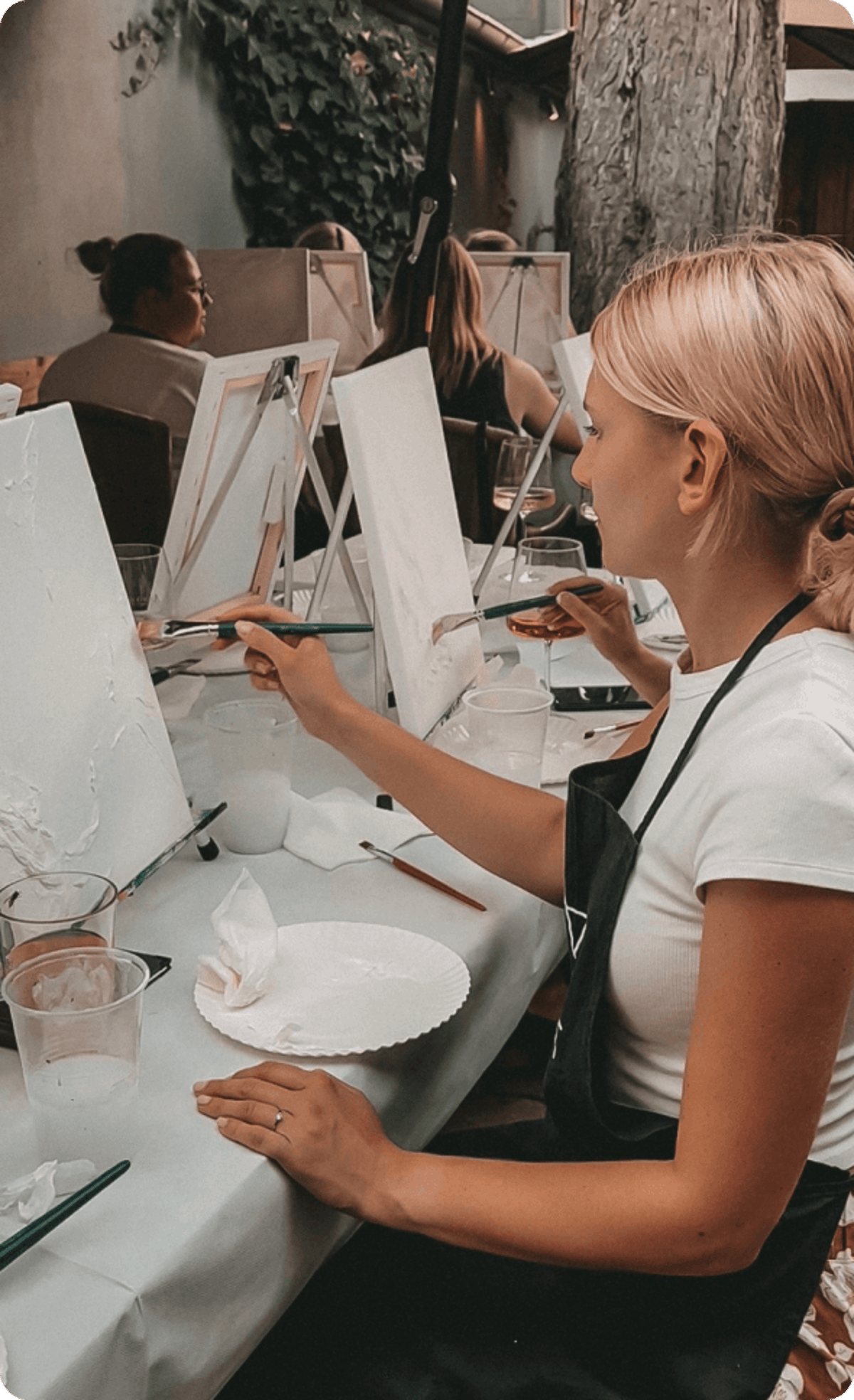 Eine Person mit blondem Haar, die in einer Gruppe auf einer Leinwand malt, hält einen Pinsel und ein Glas.