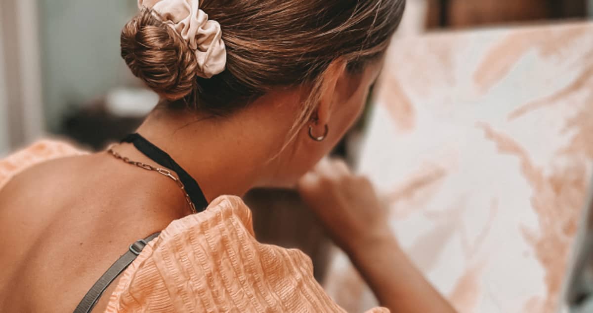 Frau mit Dutt und Haargummi malt auf einer Leinwand, trägt ein orangefarbenes strukturiertes Oberteil und ein schwarzes Band um den Hals.